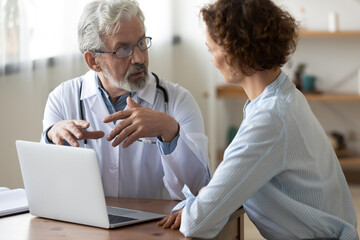 Mature Caucasian male doctor consult female patient discuss anamnesis work on computer together. Senior man GP have consultation with woman use laptop for prescription. Healthcare, medicine concept.
