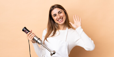Woman using hand blender over isolated background saluting with hand with happy expression
