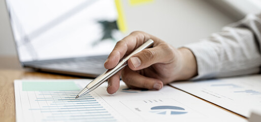 A close-up view of a businessman holding a pen pointing at a bar chart on a company financial document prepared by the Finance Department for a meeting with business partners. Financial concept.