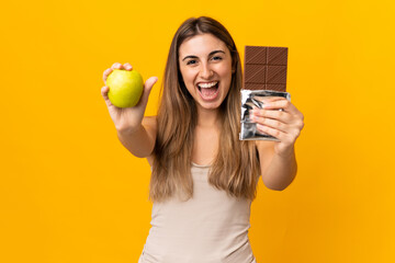 Young woman over isolated yellow background taking a chocolate tablet in one hand and an apple in...