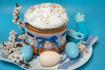 Easter composition. Eggs, apricot flowers, a cute handmade bird and a glazed Easter cake decorated with sugar sprinkles on a blue background. Happy Easter Holidays. Close-up, selective focus.