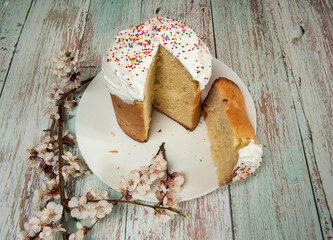 Easter composition. A branch of a blooming apricot and a glazed Easter cake decorated with sugar sprinkles, with a cut slice close-up on a white plate on a light wooden background. Selective focus.