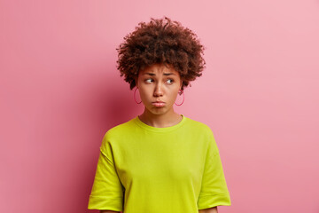 Sad gloomy young Afro American woman looks displeased aside purses lips feels upset and disappointed dressed in casual clothes isolated over pink background waits for apology expresses regret