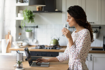 Healthy habit. Relaxed female freelancer working from home start work day from glass of pure crystal water check mail read news on laptop. Happy young woman drink mineral aqua at kitchen by pc screen.