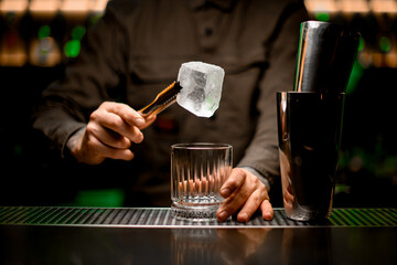 male bartender holds large piece of ice with tongs over empty glass on bar counter