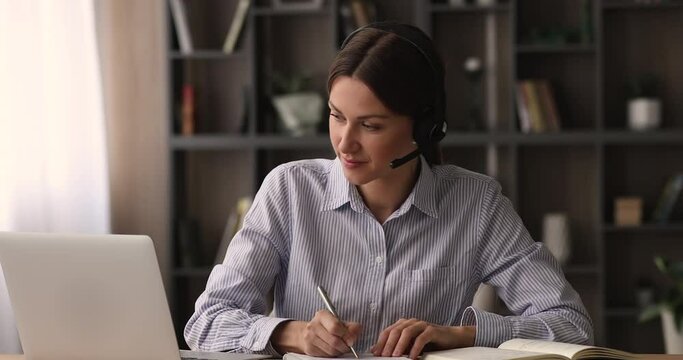 Concentrated 35s young woman in headset with mic listening educational lecture online or taking part in video call zoom meeting business negotiation, writing notes, working distantly in office or home