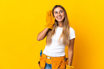 Young hispanic electrician woman over isolated yellow wall listening to something by putting hand on the ear