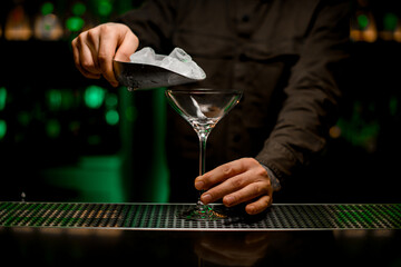 beautiful view of martini glass in which man bartender pours ice cubes from scoop