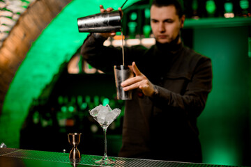 view on martini glass with ice and jigger on bar counter. Blurred bartender in background