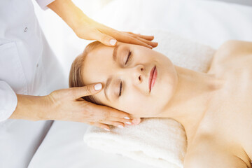 Fototapeta na wymiar Beautiful caucasian woman enjoying facial massage with closed eyes in sunny spa salon. Relaxing treatment in medicine