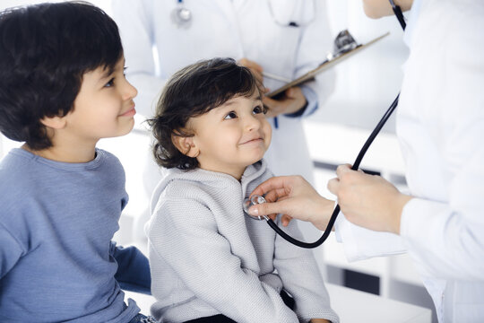 Woman-doctor examining a child patient by stethoscope. Cute arab toddler and his brother at physician appointment. Medicine concept