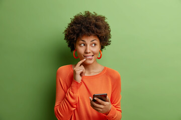 Fototapeta na wymiar Good looking dark skinned young woman with curly hair holds modern smartphone uses new application dressed in casual orange sweater isolated over green background thinks about something pleasant