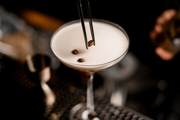 close-up of glass with frothy espresso martini cocktail decorated with coffee beans and tweezers