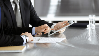 Unknown businessmen using tablet computer and work together at the glass desk in modern office, close-up. Teamwork and partnership concept