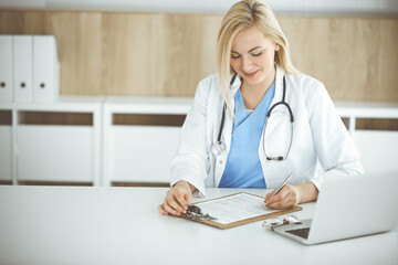 Woman-doctor at work while sitting at the desk in hospital or clinic. Blonde cheerful physician filling up medication history record form