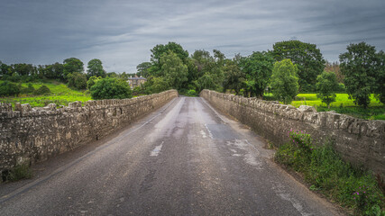 Fototapeta na wymiar Road leading over old, 12th century stone Bective Bridge over Boyne River with stone wall on sides, Count Meath, Ireland