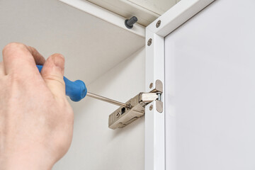 Skilled handyman adjusts furniture hinge with screwdriver installing contemporary white case in...