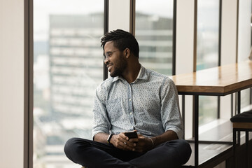 Smiling dreamy African American man wearing glasses distracted from phone, looking out window, holding smartphone, visualizing good future, waiting for call or message, enjoying leisure time