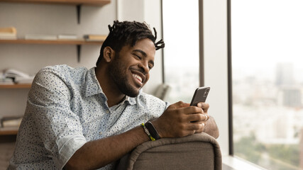 Close up smiling African American man using phone, chatting with girlfriend in social network, enjoying leisure time with mobile device, browsing apps, shopping online, sitting on couch at home