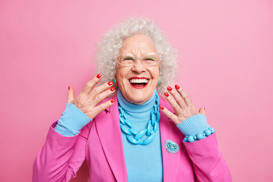 Portrait Of Elderly Curly Haired Woman Raises Hands Smiles Broadly Wears Spectacles Fashionable Outfit Necklace Being In Good Mood Isolated Over Pink Background. Old People And Style Concept