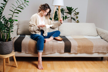 Focused young woman sewing in comforts of her apartment. She is sitting on a couch, embroidering a...