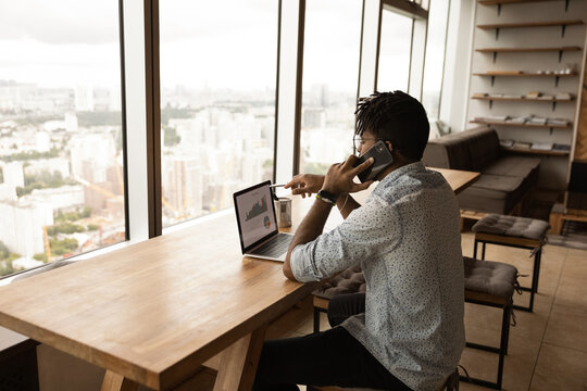 Back view African American businessman talking on phone, discussing project with colleague or business partner, using laptop, analyzing financial statistics, diagrams, broker consulting client