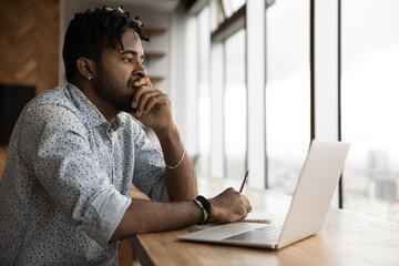 Close up thoughtful anxious African American man touching chin, looking to aside, sitting at table with laptop, pensive worried businessman pondering, solving problem, business failure, bankruptcy
