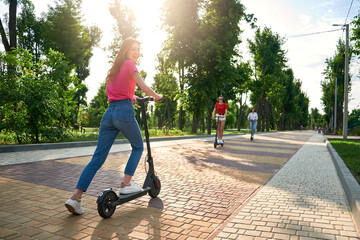 Three young girl friends on the electro scooters having fun in city street at summer sunny day. Outdoor portrait of three friends girl riding electric kick scooter in the park © irissca