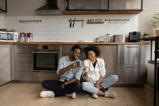 Overjoyed African American couple drinking tea, clinking cups, sitting on kitchen floor at home, happy wife and husband, satisfied customers celebrating relocation in new apartment, mortgage - Powered by Adobe