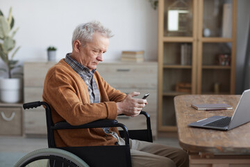 Side view portrait of senior man in wheelchair using smartphone while sitting by desk in home interior, copy space