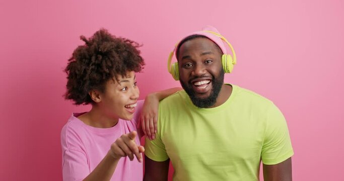 Hey look there. Positive Afro Americann woman points directly at camera shows something funny to boyfriend who listens audio track via headphones giggle together stands closely indoor over pink wall