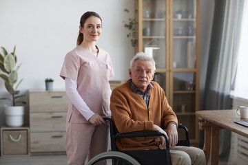 Portrait of young female nurse looking at camera while helping senior man in wheelchair, copy space