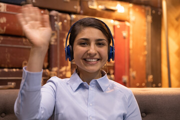 Head shot portrait smiling Indian woman wearing headphones waving hand at camera, businesswoman involved in internet meeting, video call, greeting viewers, happy mentor teacher leading online lesson