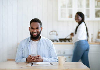 Young African Guy Sitting At Kitchen With Smartphone While Wife Cooking Lunch