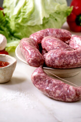 Raw uncooked italian sausages salsiccia in plate on white marble table. Green salad, vegetables and tomato sauce around.