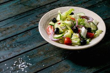 Classic vegetable salad with tomatoes, cucumber, onion, salad leaves and black olives in white ceramic plate. Dark wooden table.