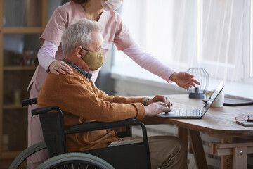 Cropped side view portrait of senior man in wheelchair using computer with nurse assisting him,...