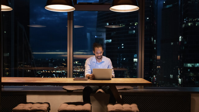 Full length African American businessman working on laptop in office at night alone, looking at screen, sitting at desk near panoramic window, employee finishing project at late hours, deadline