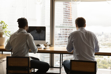 Back view diverse colleagues employees sitting at wooden desk near panoramic window in modern...
