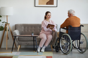 Full length portrait of smiling female nurse talking to senior man in wheelchair and using digital...