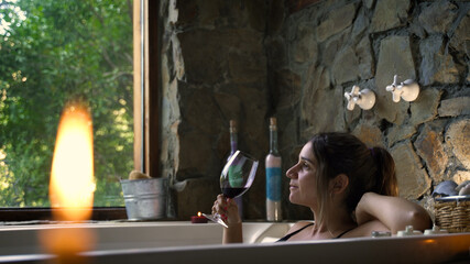 Young attractive female having a fancy bath in a hot tub spa drinking wine with big green nature...