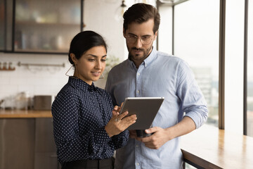 Close up diverse colleagues using tablet together, discussing online project, smiling Indian businesswoman and Caucasian businessman wearing glasses looking at device screen, standing in office