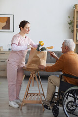 Vertical full length portrait of young female caregiver bringing groceries to senior man in...