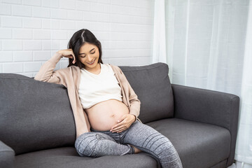 beautiful asian pregnant woman placing hands on baby lump feeling heartbeat of baby smiling joyfully, sitting on sofa relaxing resting from tiredness, living room with brick texture wall grey couch