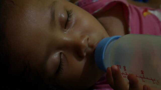 Portrait of little cute baby girl lies on bed sucks bottle of milk, drinks with big appetite.