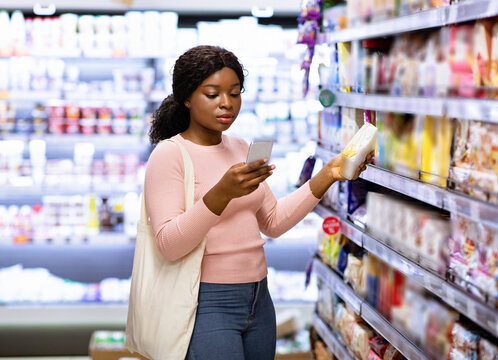 Serious black woman with eco tote bag buying groceries at supermarket, checking shopping list on smartphone