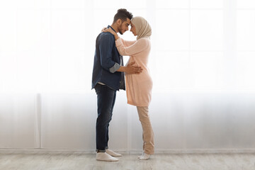 Romantic muslim spouses expecting baby standing together against window at home