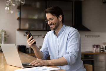 Naklejka na ściany i meble Close up smiling businessman wearing glasses using laptop and phone, looking at smartphone screen, working on online project, financial statistics, chatting with business partner, consulting