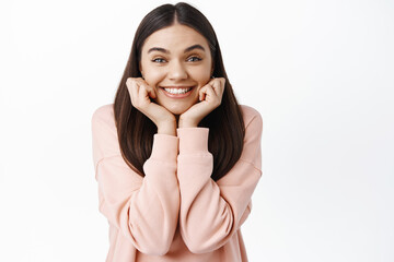 Portrait of excited smiling girl leaning on face and looking enthusiastic at camera, gazing at...