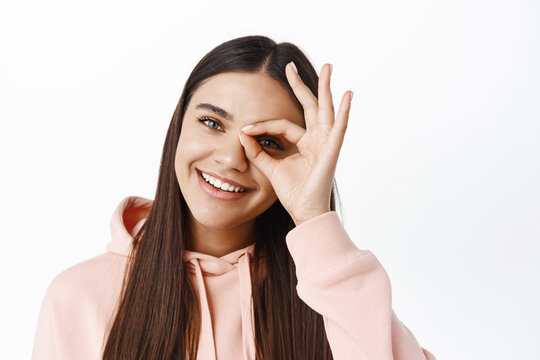 Close up of young happy woman making okay sign on eye, say yes, praise something good, standing against white background